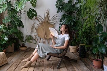 Fototapeta na wymiar Happy young caucasian woman florist relax in home garden breathing fresh air hold paper fan in hand. Relaxed carefree millennial female gardener refreshing in indoor orangery during hot summer day