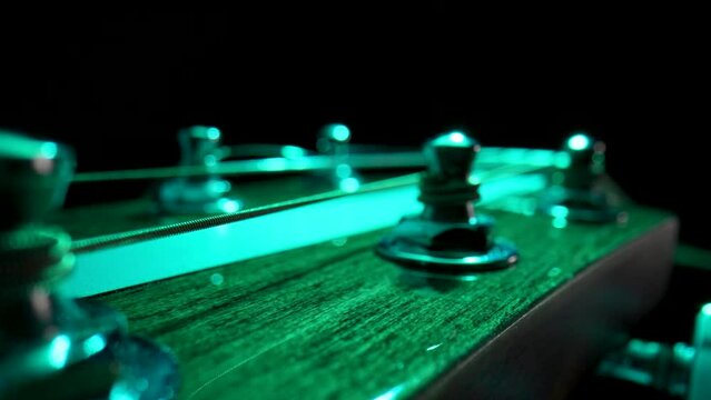 Wooden guitar's headstock with metal tuning pegs and string illuminated by green light on black background. Stringed musical instrument with pegs for tensioning and tuning sound of strings. Close up.
