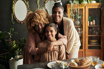 Portrait of three sisters embracing and smiling at camera while sitting at table during family...