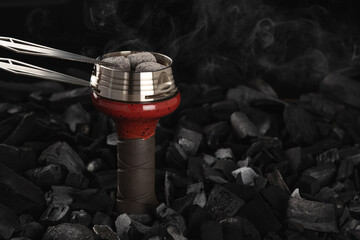 Metal kaloud with red-hot cubes of coconut charcoal with tongs is placed on a ceramic bowl for a...