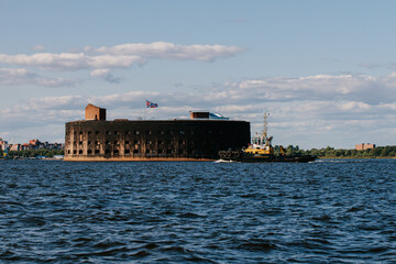 Fototapeta premium View from a boat on Fort Alexander the First in Kronstadt in the waters of the Gulf of Finland. A tugboat ship sails in the distance. Summer day