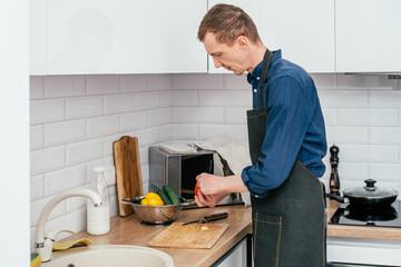 Portrait of middle-aged man wearing blue long-sleeve shirt and black apron chopping red bell pepper with knife on wooden cutting board on kitchen table. Cooking, healthy food.