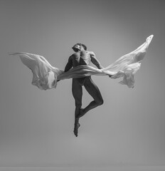 Flight. Black and white portrait of graceful muscled male ballet dancer dancing with fabric, cloth isolated on grey studio background. Grace, art, beauty concept. Weightless, flexible.