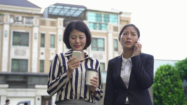 employee in causal smart is phubbing with a smile while waiting for her coworker who&rsquo;s having a business talk on the smartphone near their company.
