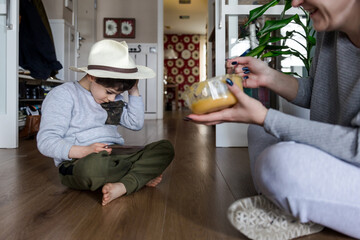 Mother feeding a boy with autism wearing a hat, sitting on the floor while playing with the mobile phone