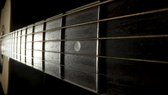 Camera pans over the fretboard of a classical acoustic guitar against a black background. Brown wooden guitar neck with metal strings and frets extreme close up.