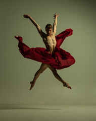 Flight. One young muscled man, flexible ballet dancer in action with red fabric, cloth isolated on olive color background. Theater, emotions, grace, art, beauty concept.