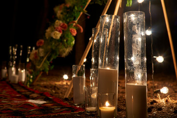 Candles burning in glass vases, flasks on carpet near bohemian tipi arch decorated with roses and flowers wrapped in fairy lights on outdoor wedding ceremony venue in pine forest. Boho rustic decor. © artiemedvedev