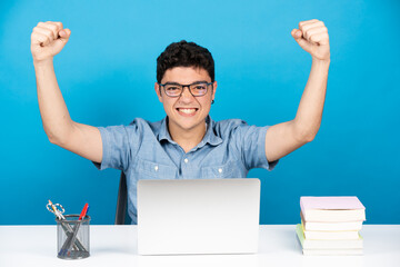 Hispanic teenager boy celebrating with arms up and looking at camera in front of laptop isolated on...