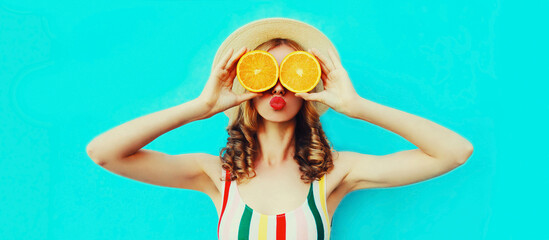 Summer portrait of cheerful young woman covering her eyes with slices of orange fruits and looking for something wearing straw hat on blue background