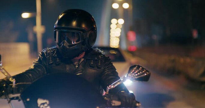 Rock Biker is Riding fast on City streets at night turning left on crossroads. Motorcyclist in black helmet drives Motorbike in evening with lights. Nightlife Urban Traffic. 4K close up tracking shot