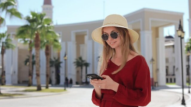 Happy caucasian woman using modern smartphone while standing among Land of Legends theme park. Beautiful lady wearing sunglasses, straw hat and red summer dress.