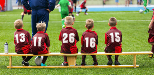 School soccer team witch coach on tournament match sitting on wooden bench. Children in red soccer shirts ready to play football game