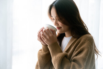 Young asian woman standing beside window and holding mug in bedroom at home, She drinking milk after wake up in the morning