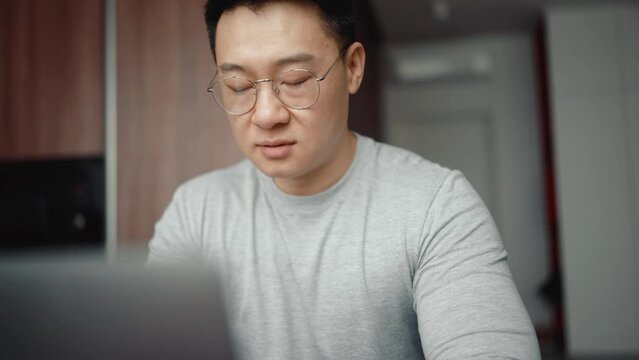 Asian happy man wearing eyeglasses typing on laptop at home