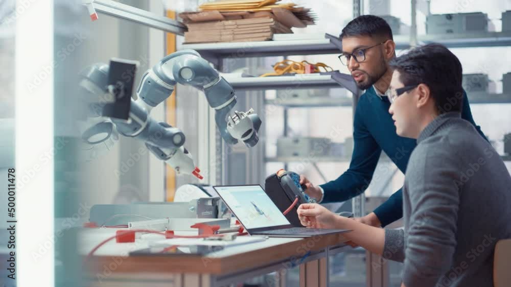Poster Black Male Engineer Tell his Ideas to Asian Developer while Control Robot Arm at Factory Workshop. People Designing Automated Robotic Systems and Startup Concept