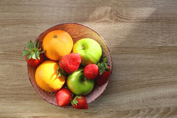 Pink bowl filled with fresh apples, oranges and strawberries on wooden table. Flat lay.