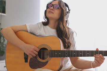 A young blogger girl is sitting on the bed playing guitar, broadcasting her performance via the...