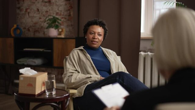 Young female patient is talking to psychoanalyst and sitting in chair in modern office spbas. 4k American African woman tells story and looks with smile, experienced specialist gives advice and sits