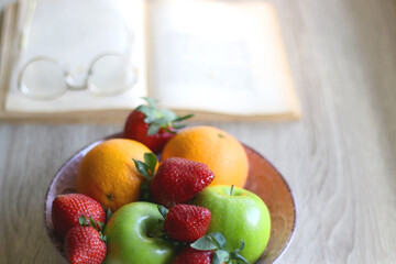 Bowl of apples, oranges and strawberries, open book and reading glasses on the table. Selective focus.