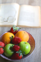 Bowl of apples, oranges and strawberries, open book and reading glasses on the table. Selective focus.