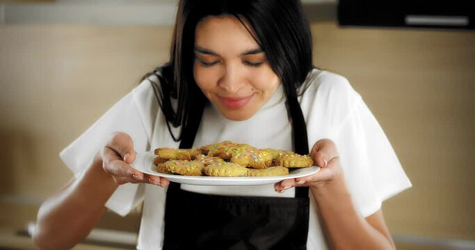 Pretty woman mixed-race brunette dressed in a white T-shirt and a brown apron sprinkles decorations on shortbread cookies and gingerbread lying on parchment on a baking sheet
