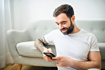 portrait of casual young man with beard and tattoo and white shirt sit on the sofa at home with...