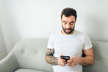 portrait of casual young man with beard and tattoo and white shirt sit on the sofa at home with...