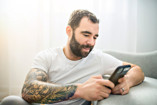 Portrait Of Casual Young Man With Beard And Tattoo And White Shirt Sit On The Sofa At Home With Cellphone