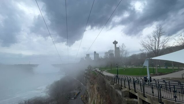 The Niagara falls, Ontario city skyline on background, under a mist cloudy foggy day from waterfall. Panning left with zip lines overhead, Niagara river on left, and tall buildings and hotels on right