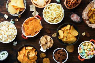 Unhealthy food or snacks. All classic potato snacks with peanuts, popcorn and onion rings and salted pretzels in bowl plates on old wooden background. Unhealthy food for figure, heart, skin, teeth.
