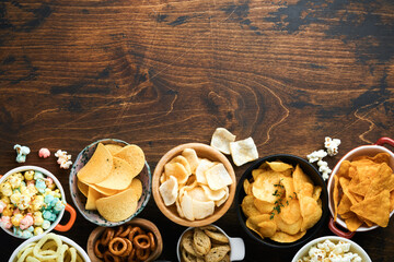 Unhealthy food or snacks. All classic potato snacks with peanuts, popcorn and onion rings and salted pretzels in bowl plates on old wooden background. Unhealthy food for figure, heart, skin, teeth.