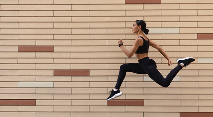 Serious young arabic female athlete in sportswear runs, frozen in air on brick wall background,...