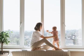 Mother and little daughter sitting at home on a windowsill by a big window, looking outside, on a high floor.