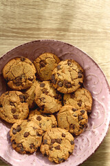 Pink plate filled with chocolate chip cookies on wooden table. Flat lay.