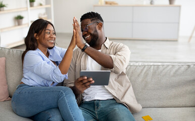 Happy black couple using tablet, celebrating win giving high five