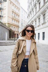 Obraz premium pleased young woman in trendy sunglasses and trench coat walking on street of vienna.