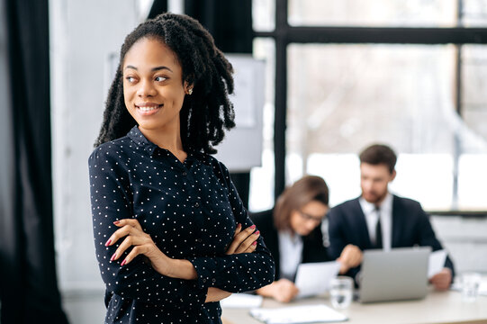 Portrait of lovely elegant young business woman at the briefing meeting. Successful african american female, stands in creative modern office with arms crossed, looks looks to the side, smile,thinking