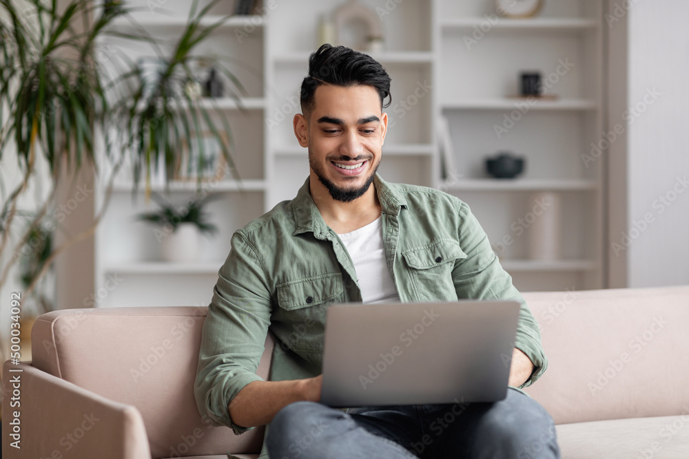 Wall mural happy attractive millennial arabic male with beard in casual sits on sofa, typing on laptop in livin