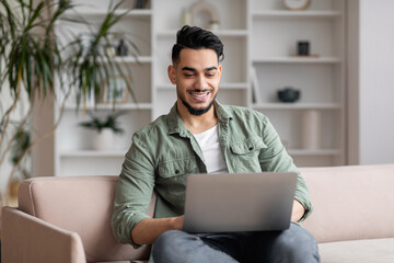 Happy attractive millennial arabic male with beard in casual sits on sofa, typing on laptop in...