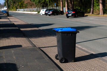 A black trash can with a blue lid on the edge of the street. Parked cars.