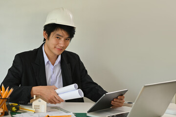 A portrait of an architecture wearing a helmet working on a tablet, laptop and tools in the office, for architect, business, safety and technology concept.