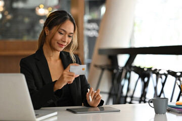 A portrait of a pretty Asian businesswoman sitting in a workplace holding a credit card and the other hand using a tablet and laptop, for business, finance and technology concept.