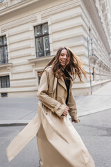Fototapeta premium happy young woman in beige trench coat walking on european street.
