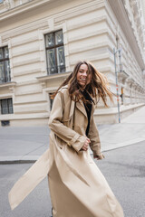 happy young woman in beige trench coat walking on european street.