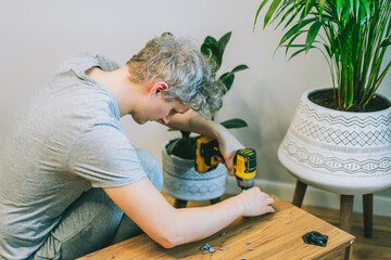 A young man with an electrical screwdriver assembles a tv stand console according to instructions in his new house. Man assembling furniture at home using a cordless screwdriver. Selective focus.