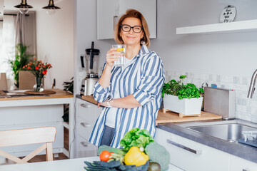 Healthy habit to drink water. Smiling middle age woman with glass of pure water with lemon standing on her kitchen. Control body hydration, Track water balance. Healthy living. Selectivbe focus. - Powered by Adobe