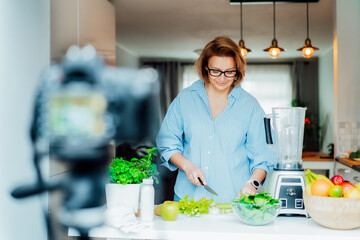 Middle age woman is blogging for her wellness lifestyle channel about healthy balanced living in the kitchen of her home. Creating video content for social media with camera on a tripod. - obrazy, fototapety, plakaty
