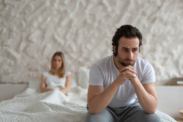Young Caucasian man feeling upset, sitting on bed at home, not speaking to his offended wife after...