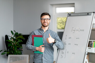 Positive mature male teacher showing thumb up standing near blackboard, recommending new online...